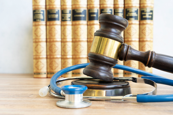 A gavel rests on a judge's block next to a stethoscope, with a row of law books in the background, symbolizing the intersection of law and medicine.